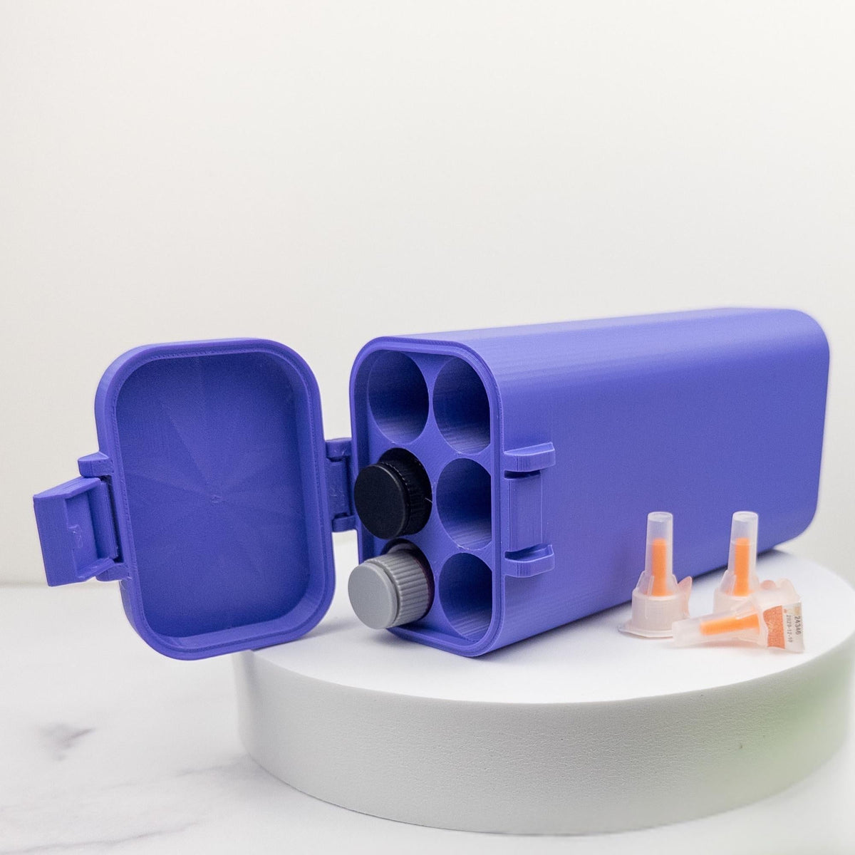A purple plastic container with a lid and two orange syringes are placed on a white surface.