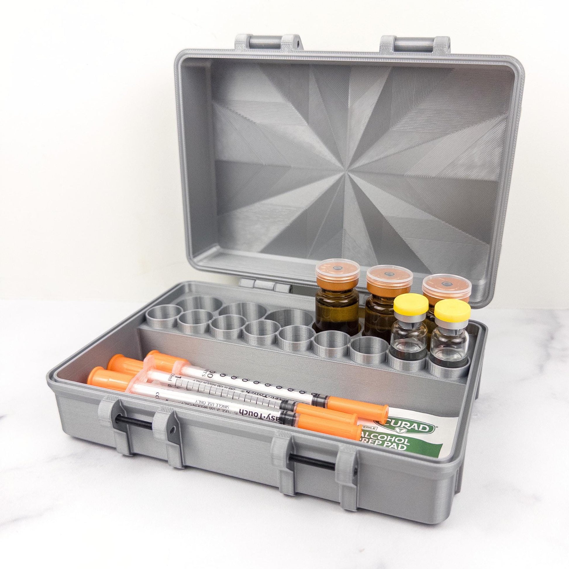 May include: A gray, open medical case with a geometric pattern on the lid. Inside are vials, syringes, and alcohol prep pads. The case has a handle and latches for secure closure. The syringes have orange plungers.